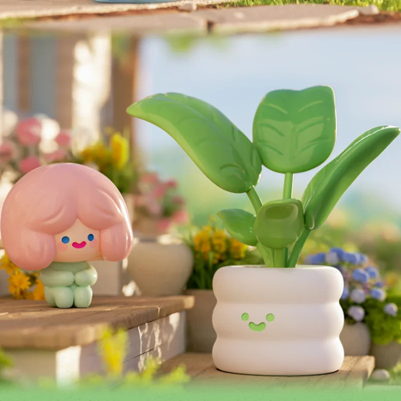 Cute figurine with pink hair and green body next to a potted plant with a white pot on a blurred outdoor background.