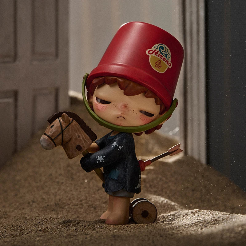 Toy figure of a child with a red bucket on its head, holding a horse and an arrow, standing in sand.