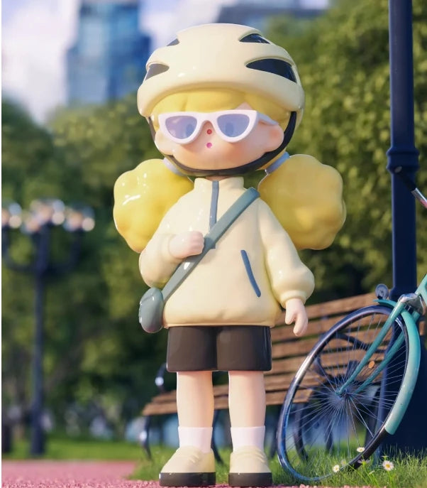 Toy figure of a person wearing a helmet and sunglasses, standing in a park-like setting with a bicycle and bench in the background.