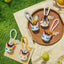 Cat-shaped keychains on a woven mat with grass and fruits in the background
