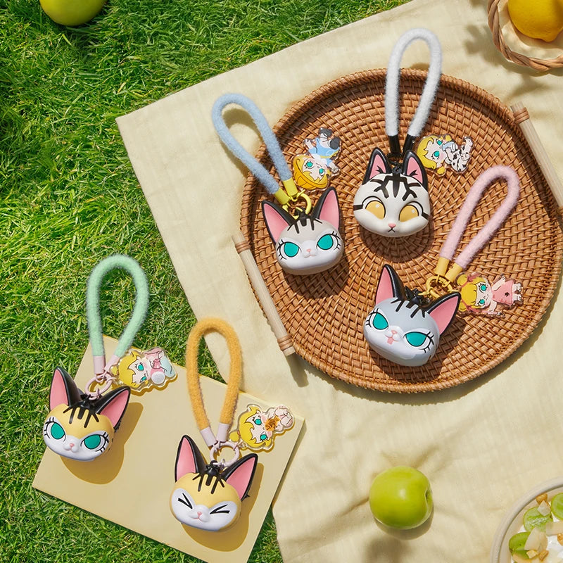 Cat-shaped keychains on a woven mat with grass and fruits in the background