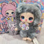 Small doll with gray fur outfit and colorful buttons, standing in front of a colorful background with cartoon characters.