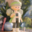 Figurine of a person in athletic wear with a green headband and bag, standing indoors with plants in the background.