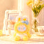 Yellow plush toy with blue bow on a decorative table with flowers and a photo frame.
