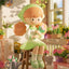 Plush toy of a girl in a green dress sitting on a stool with flowers in a garden setting.