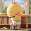 Toy figure with blonde hair and a book, sitting on a rug with a blurred bookshelf in the background.